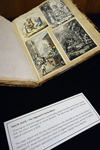Open album on display, showing four drawings or paintings pasted onto the righthand page, with descriptive text card visible at bottom of image.